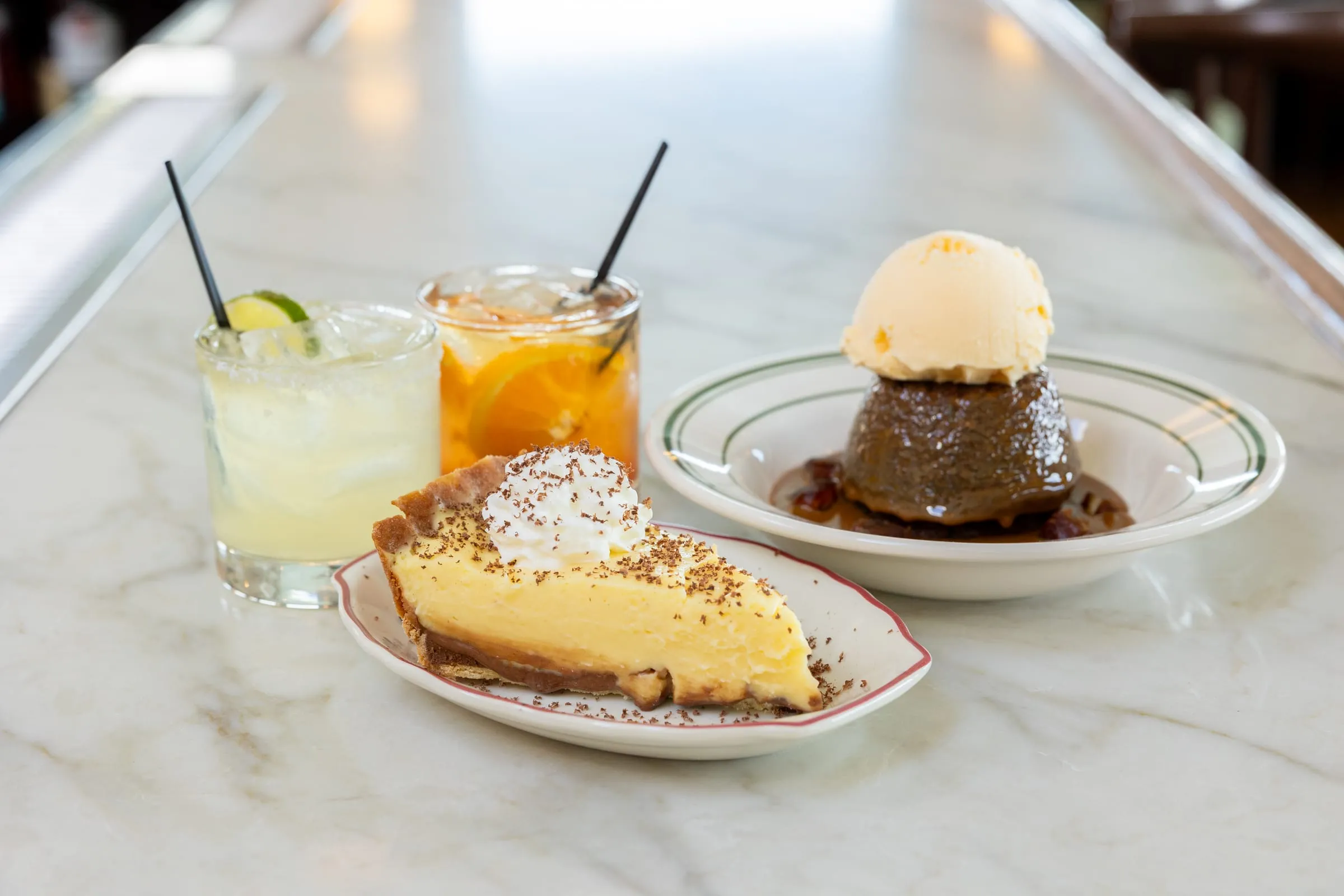 A slice of fudge bottom pie, sticky toffee pudding topped with ice cream, and two cocktails on the marble bar top.