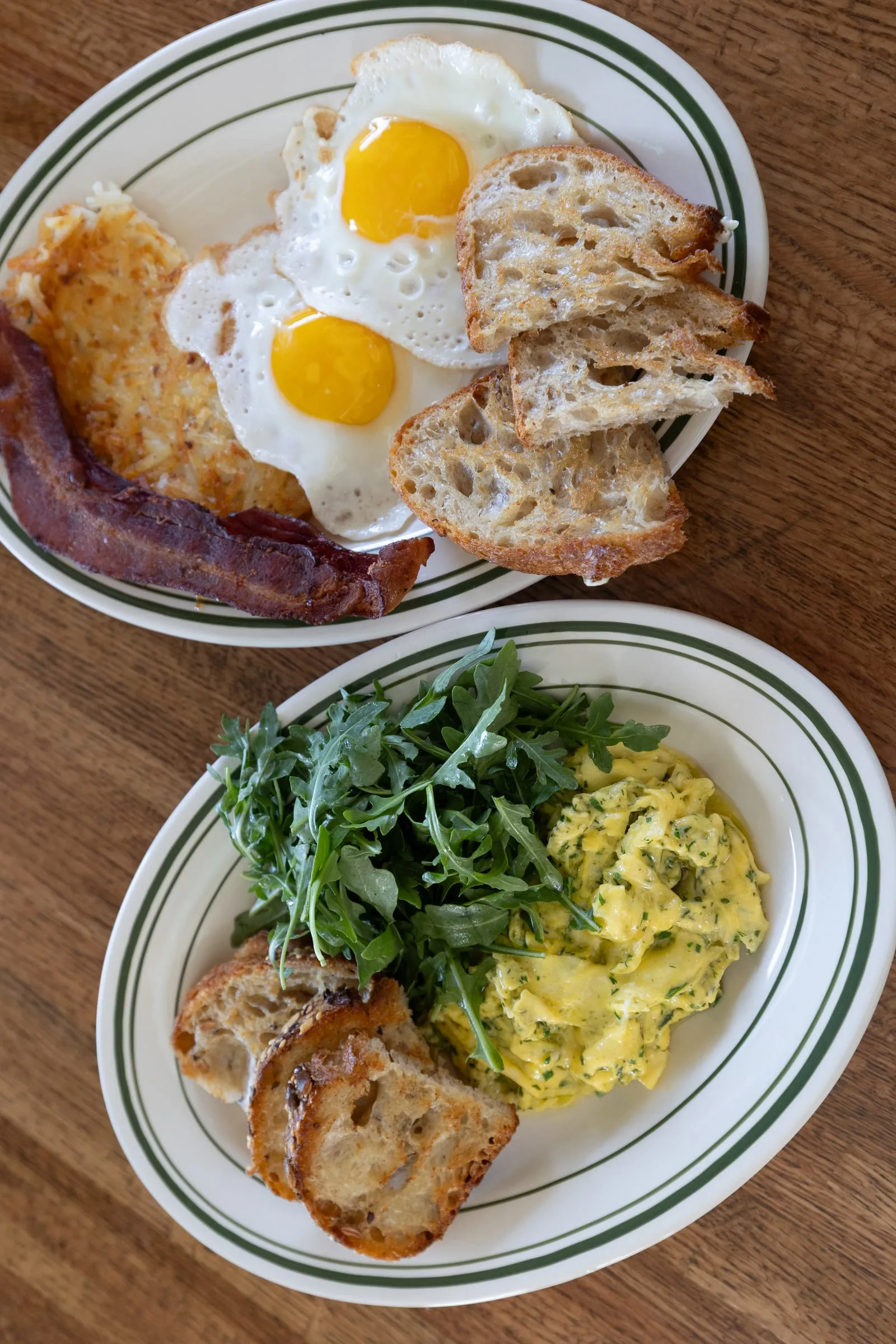 Two sunny-side-up eggs with bacon, hash browns, and sourdough toast beside a plate of herb scrambled eggs with arugula.