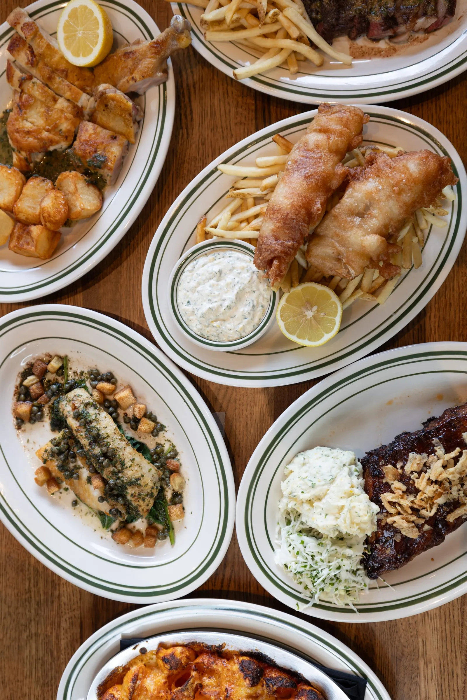A spread of Emmi's entrées on green-striped plates: walleye fish and chips with tartar sauce, roast chicken with crispy potatoes, ribs with slaw and potato salad, and steak frites.