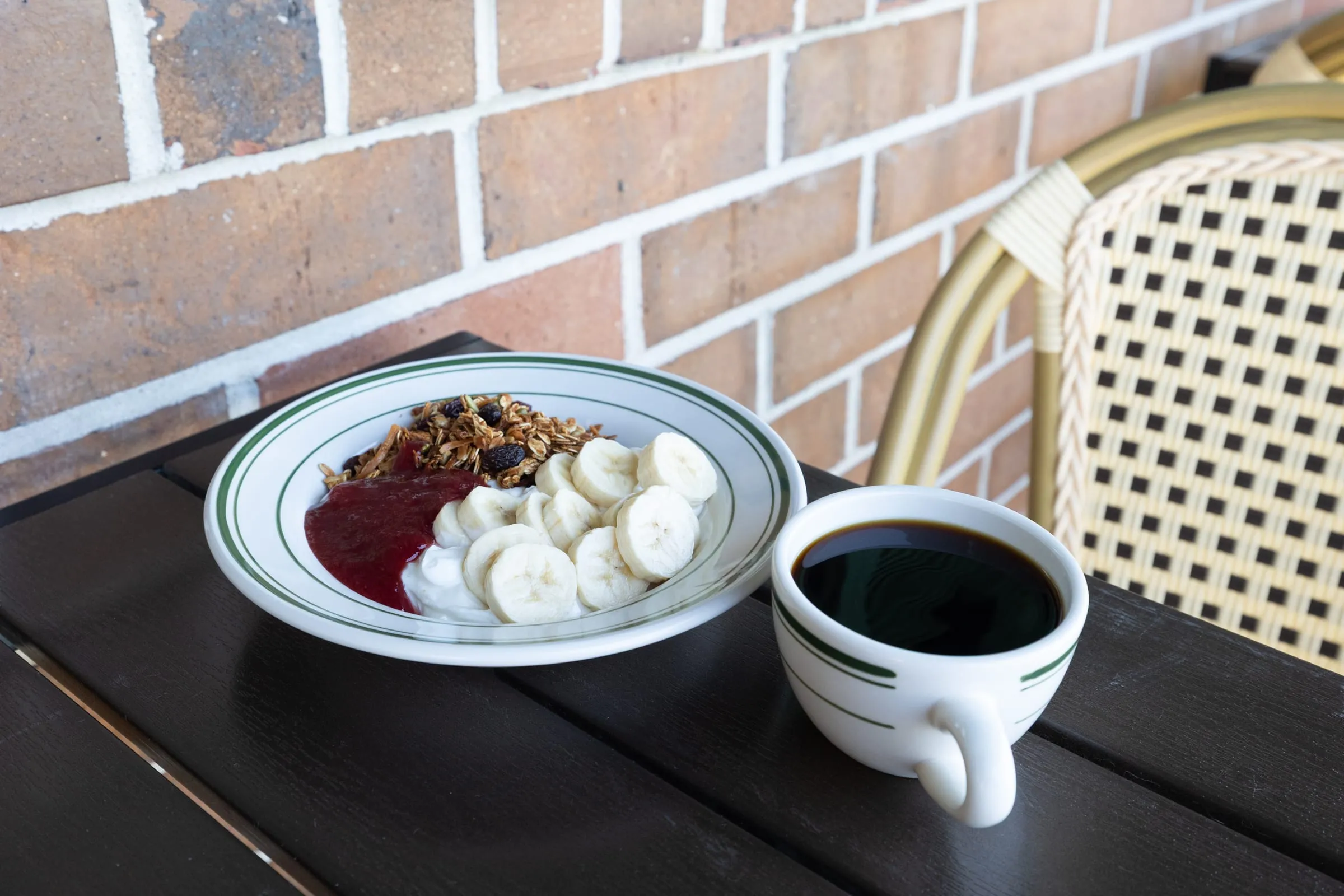 A cup of black coffee in a green-striped diner mug beside a bowl of yogurt with granola, bananas, and berry compote on the outdoor deck.