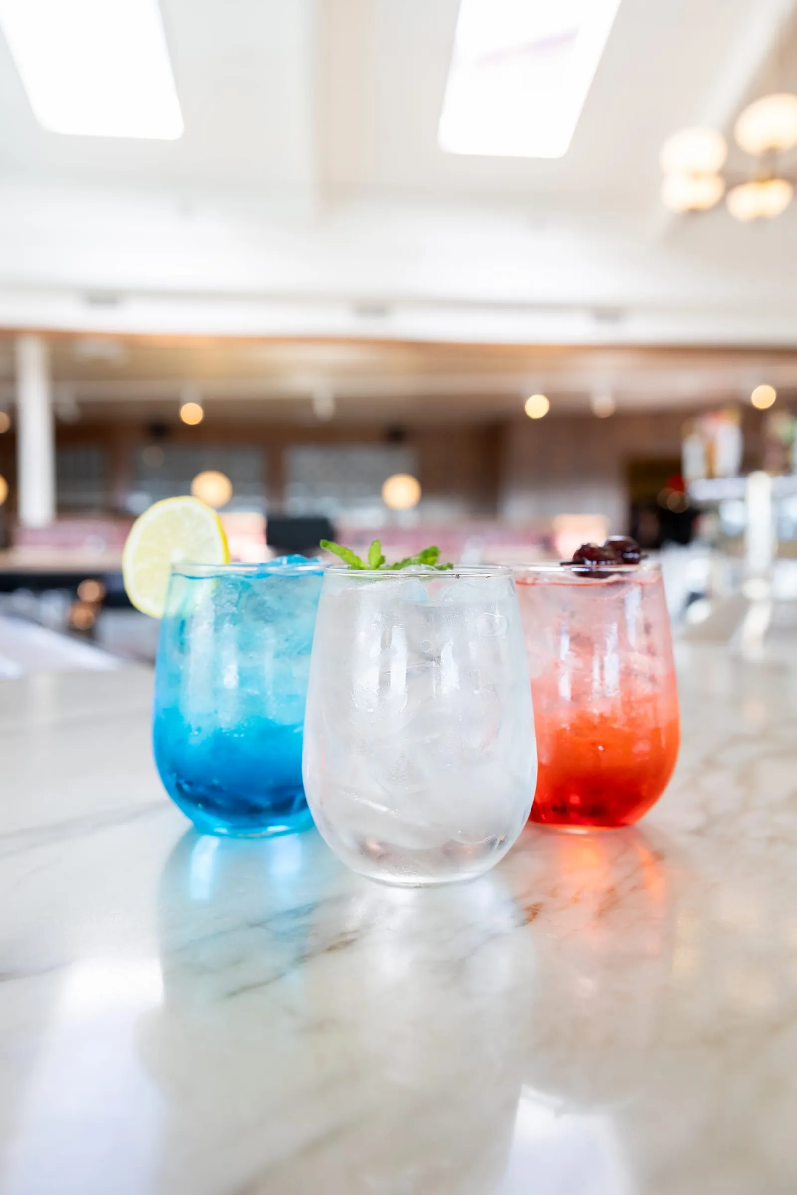 Three cocktails in red, white, and blue lined up on the marble bar, garnished with lemon, mint, and cherries.